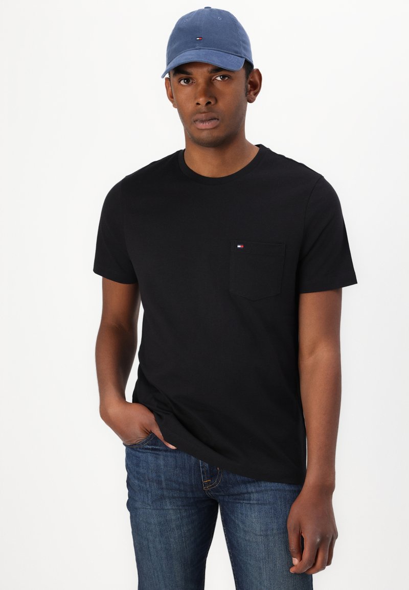 Czarna koszulka z krótkim rękawem z kieszonką i małym logo, w zestawieniu z niebieskimi dżinsami i jasnoniebieską czapką. Gładki materiał, luźny fason.