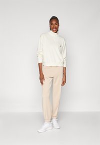 Lichte crème sweatshirt met gedeeltelijke rits en logopatch, gecombineerd met getailleerde lichtbeige joggers en witte sneakers. Glad stoftextuur.