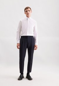 Camicia bianca a maniche lunghe, abbottonata, abbinata a pantaloni sartoriali blu scuro. Il modello sta in posizione formale, indossando scarpe di pelle nere. Sfondo semplice.