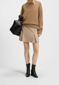 Pull en maille marron, jupe plissée beige à fines rayures, et bottines noires à talons. Sac noir avec poignée plate.