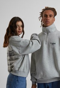 Dos personas llevan sudaderas grises. Una tiene un cuello alto con cremallera; la otra tiene mangas anchas y sueltas con texto en la parte trasera. Ambas son informales.