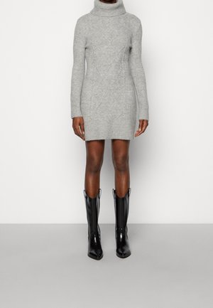 Femme portant une robe-pull à col roulé gris clair en maille torsadée et des bottes de cowboy noires brillantes mi-mollet, debout devant un fond uni.