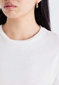 Witte, korte-sleeve shirt met een ronde halslijn, gladde textuur. Model draagt een gouden ringoorbel, zichtbaar van de schouders omhoog.