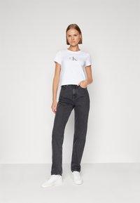 Camiseta blanca de manga corta con un logo "CK" gris, combinada con pantalones vaqueros oscuros de talle alto y zapatillas blancas. Fondo liso.