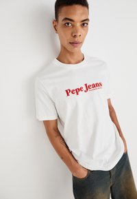 Balts īsām piedurknēm T-krekls ar izteiktu sarkanu "Pepe Jeans" logo priekšpusē. Gluda auduma, klasiskā apaļā kakla dizains, brīvs griezums.