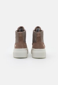 Een paar bruine suède en stoffen hoge sneakers met witte dikke zolen, gezien vanaf de achterkant op een effen witte achtergrond.
