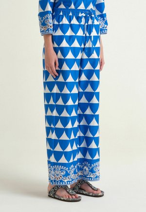 Femme portant un pantalon bleu et blanc à motif géométrique avec une bordure florale et des sandales ouvertes décorées.