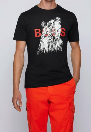 Mann trägt ein schwarzes T-Shirt mit einer weißen Skizze eines Bärenkopfs und rotem Schriftzug "BEARS", kombiniert mit leuchtend orangefarbenen Cargo-Hosen.