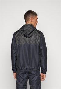 Rompevientos de la marina con capucha; tela de patrón geométrico de color verde oscuro en la sección superior; puños y cintura elásticos; textura suave.