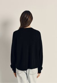 Črn pleten sveterek z ohlapnim krojem, okroglim izrezom in dolgimi rokavi, v kombinaciji s svetlo modrimi kavbojkami. Mehak material, preprost dizajn.