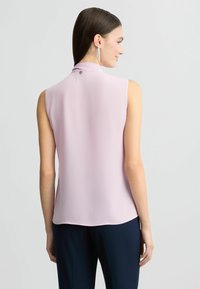 Mouwloze, lichtroze blouse met een hoge hals en een knoopsluiting aan de achterkant, gemaakt van een gladde, voortvloeiende stof. Randen zijn afgerond.