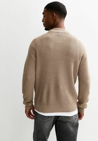 Beige gebreide trui met ribbelstructuur, ronde halslijn en lange mouwen, gestyled over een witte shirt, gedragen met donkere jeans.