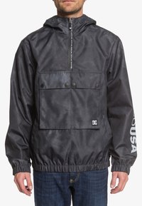 Black hooded pullover jacket in lightweight, water-resistant material. Features a zippered front, large chest pocket, and elasticized hem.