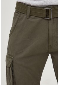 Close-up of olive green cargo pants with side pocket flap and matching fabric belt with metal buckle.