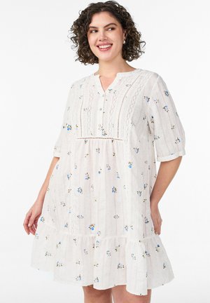 Smilende kvinde med krøllet hår iført hvid kortærmet kjole med blå blomsterprint og blonde detaljer, stående mod en hvid baggrund.