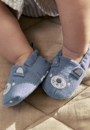 Babyben iført bløde blå tøfler med bjørneansigter, hvilende på en beige quiltet overflade.