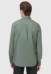 Groen overhemd met lange mouwen en kraag, met een detail van een rugplooitje en knoopsels bij de manchetten. Gemaakt van zachte stof met een gladde textuur.