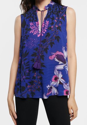 Sleeveless blue blouse with pink and black floral pattern, gathered neckline with tie and tassels, worn with black pants.