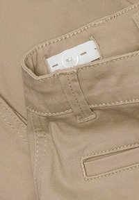 Ceinture en tissu beige avec une étiquette blanche et un bouton. Détails de couture visibles. Texture douce avec un design minimaliste de poche sur le côté droit.