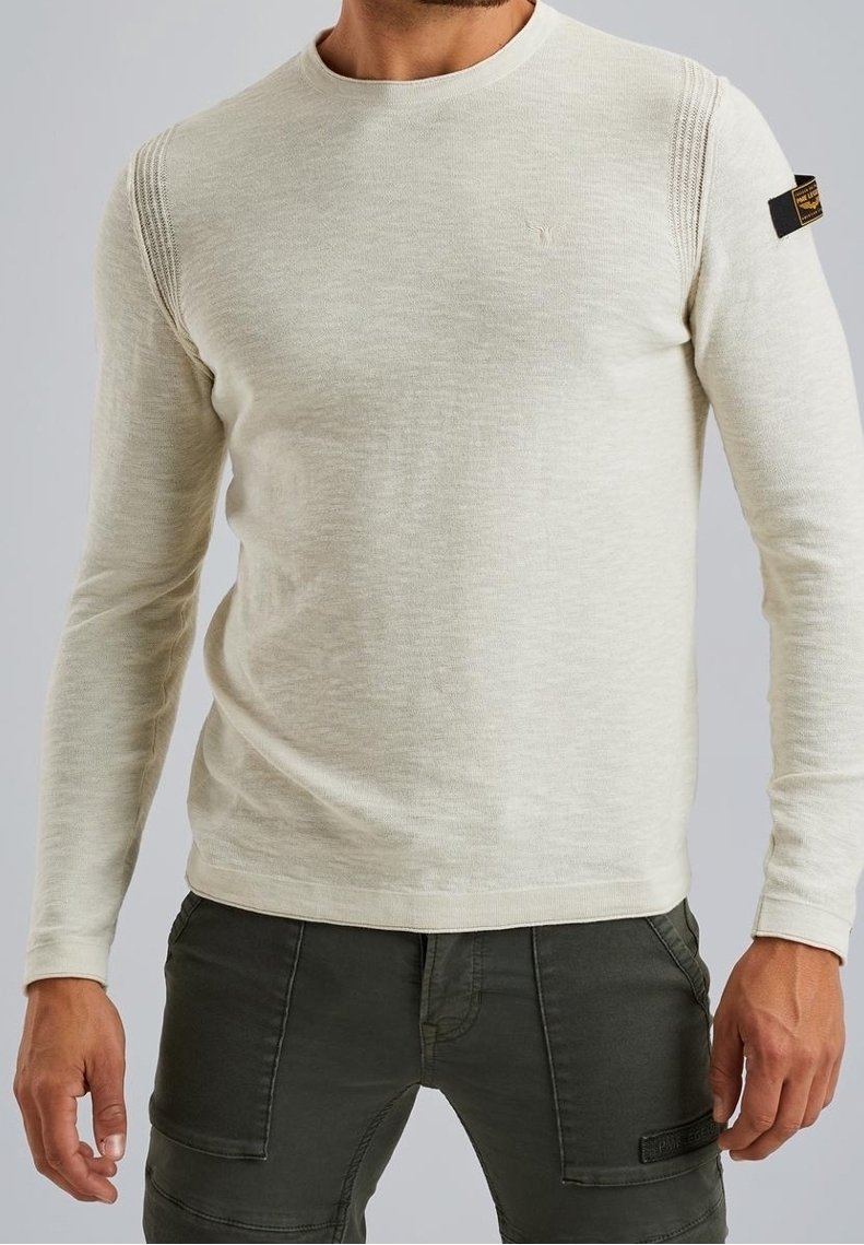 Pull à manches longues beige clair avec un col rond, des accents côtelés sur les épaules et un petit logo sur le devant, associé à un pantalon vert foncé.