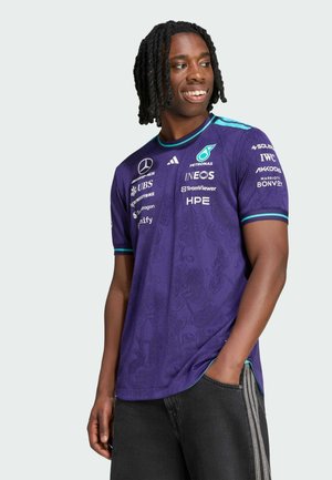 Joven con el cabello trenzado que lleva una camiseta morada de Mercedes-AMG Petronas Formula One, sonriendo y mirando hacia un lado con una mano en el bolsillo.