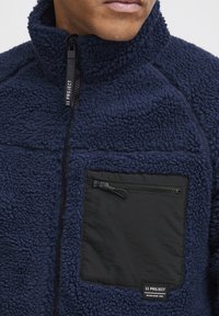 Navy fleece jacket with a zippered black chest pocket. High collar and textured fabric create a warm, layered design. Features branded tag.