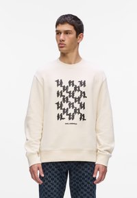 Sudadera crema con estampado gráfico negro, cuello redondo y puños acanalados. Presenta un diseño de logo repetido, corte relajado y textura suave.