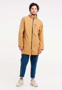 Chaqueta a prueba de agua de color miel con cremallera completa, capucha grande, bolsillo frontal con cremallera y detalles en negro, combinada con jeans azules y zapatillas blancas.