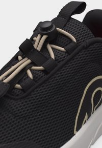 Κοντινό πλάνο μαύρου πλεκτού αθλητικού παπουτσιού με μπεζ ελαστικά κορδόνια και μαύρο κούμπωμα κορδονιού, με μπεζ και μαύρο σχέδιο στο πλάι.