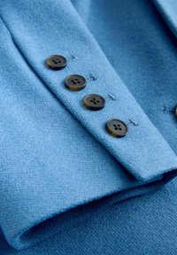 Hellblauer Wollstoff mit strukturierter Oberfläche; verfügt über vier dunkle Knöpfe entlang der Manschette und kontrastierende Naht mit Garn.