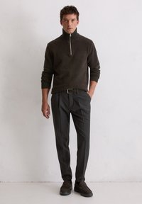 Dunkelbrauner Zip-Pullover mit strukturierter Oberfläche, kombiniert mit schwarzen, taillierten Hosen und dunklen Wildlederschuhen. Schlichte, moderne Schnittführung und Design.