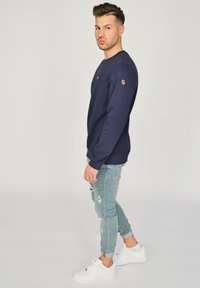 Granatowy sweatshirt z okrągłym dekoltem i ściągaczami na rękawach, w połączeniu z jasnoniebieskimi spodniami dżinsowymi z efektem przetarcia i białymi sneakersami. Logo na piersi.