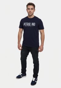 Marineblauwe t-shirt met korte mouwen met "PETROL IND" bedrukt in een versleten witte lettertype. Gekenmerkt met donkere jeans en marineblauwe sportschoenen.
