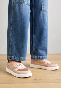 Ljusrosa sneakers med slät yta, perforerad tå och vit gummisula. Kombinerade med löst sittande blå denimjeans.