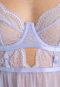 Bralet din dantelă lavandă cu motive florale, plasă transparentă și tiv din satin. Prezintă margini zimțate și detalii decupate în centrul spatelui.