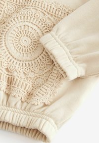 Beige garment featuring intricate circular embroidery detail in the center, gathered sleeves, and a soft texture.