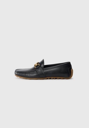 Mocassin en cuir noir à bout rond, avec une boucle décorative en or et une semelle en caoutchouc. Présente des détails de couture sur la tige et sur le côté.