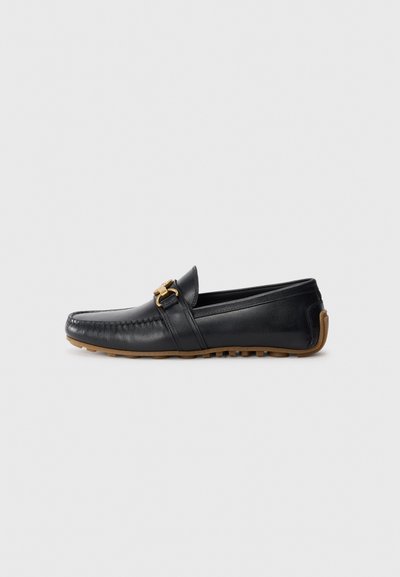 Mocasin de cuero negro con punta redonda, hebilla decorativa dorada y suela de goma. Presenta detalles de costura en la parte superior y en el lateral.