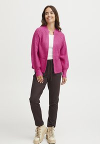 Bright pink knitted cardigan with a round neck, two front pockets, and ribbed cuffs, paired with dark pants and beige lace-up boots.