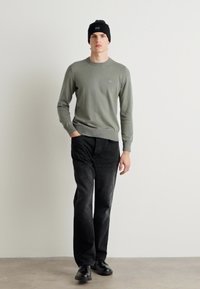 Groene gebreide trui met een ronde halslijn, gecombineerd met zwarte denim broek en een zwarte beanie. De pasvorm is ontspannen en de stof lijkt zacht te zijn.