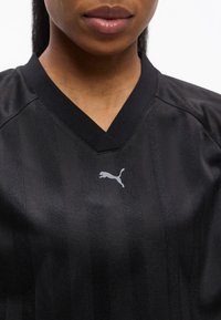 Κοντινό πλάνο ενός ατόμου που φοράει μαύρο αθλητικό μπλουζάκι Puma με V-λαιμόκοψη και διακριτικό ριγέ μοτίβο.