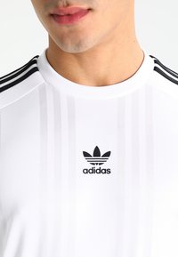 White Adidas athletic shirt featuring horizontal stripes, a ribbed crew neckline, and a black logo on the left chest.