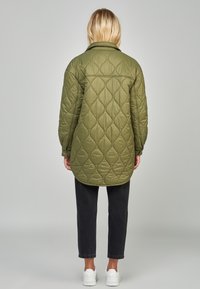 Chaqueta acolchada verde con una silueta larga, dobladillo redondeado y un patrón de diamantes texturizado, combinada con pantalones negros ajustados y zapatillas blancas.
