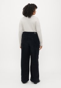 Pantaloni largi din catifea neagră cu talie înaltă, buzunare la spate și o textură netedă. Purtați cu un top ajustat de culoare deschisă.