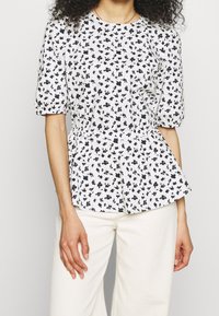 Hvid peplum-top med marineblåt blomsterdesign, korte puffærmer, rund halsudskæring og sammensnørende talje. Letvægtsstof med en tekstureret overflade.