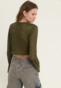 Olivgrön, kortärmad långärmad topp i mesh-material, som visar små hål för andningsförmåga. Matchad med ljusblå högmidjade jeans.