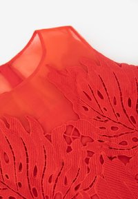 Vestido rojo sin mangas con intrincado bordado de hojas en una tela texturizada, con un escote transparente y recortes detallados.