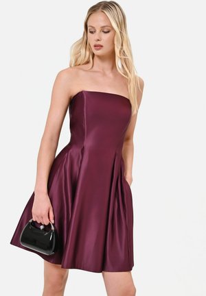 Femme portant une robe bustier brillante violet foncé, tenant un petit sac à main noir, regardant légèrement sur le côté.