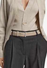 Kvinna iförd en beige strukturerad blazer och en knapplös cardigan instoppad i högmidjade mörka veckade byxor med ett flätat beige bälte.