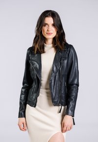 Schwarze Leder-Bikerjacke mit asymmetrischem Reißverschluss, auffälligen Revers und strukturierter Oberfläche, getragen über einem cremefarbenen gerippten Kleid.